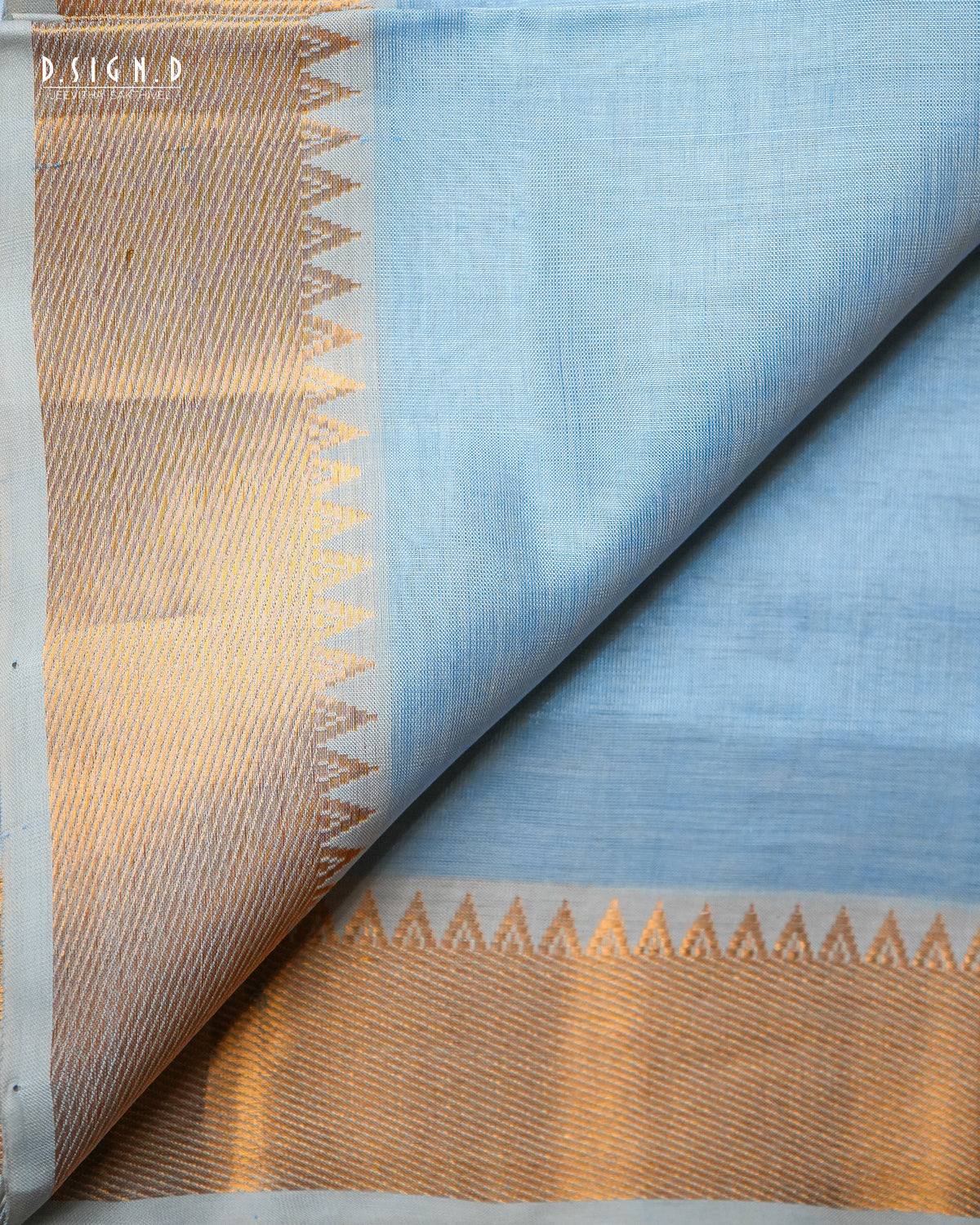 A folded light blue silk dhoti & vastram with a wide gold zari border featuring a geometric chevron pattern, displayed on a white surface.