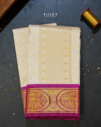 A folded cream-colored silk dhoti with a wide magenta and gold zari border, displayed on a dark textured surface next to a spool of gold thread.