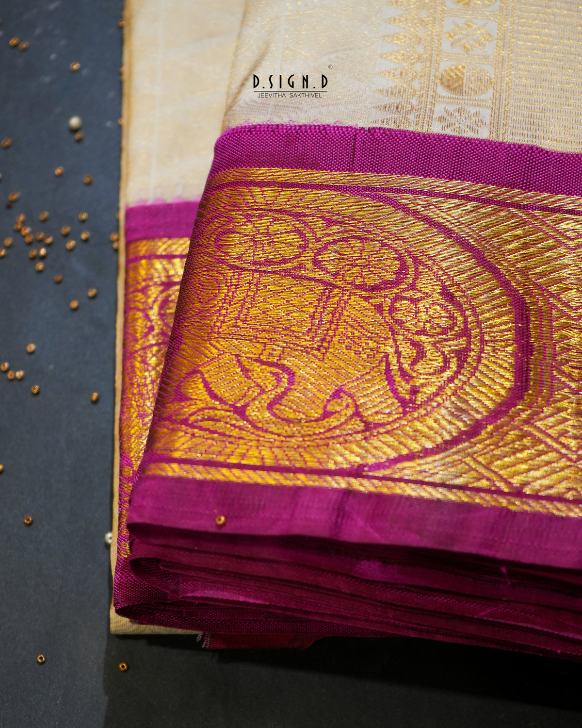 A folded Indian silk dhoti & vastram with a cream body and a rich magenta and gold zari border, displayed on a dark textured surface with scattered mustard seeds.