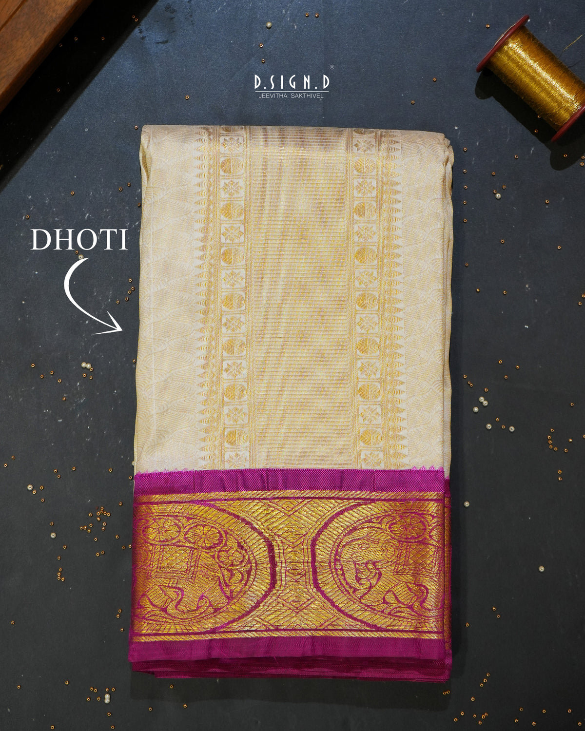 A folded cream-colored silk dhoti with a wide gold zari border and a magenta-pink pallu, displayed on a dark textured surface with 'DHOTI' text overlay.