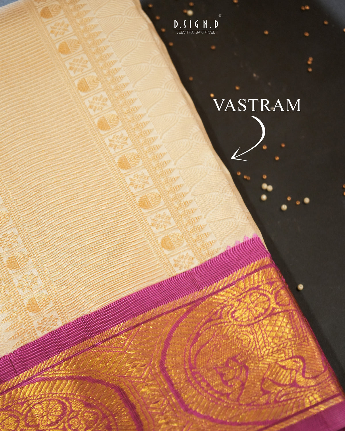 A close-up of a cream-colored silk vastram with a rich gold and magenta zari border, laid diagonally on a dark textured background with decorative seeds. The text 'VASTRAM' points to the traditional four-corner design of the border.