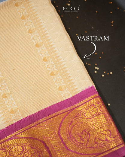 A close-up of a cream-colored silk vastram with a rich gold and magenta zari border, laid diagonally on a dark textured background with decorative seeds. The text 'VASTRAM' points to the traditional four-corner design of the border.