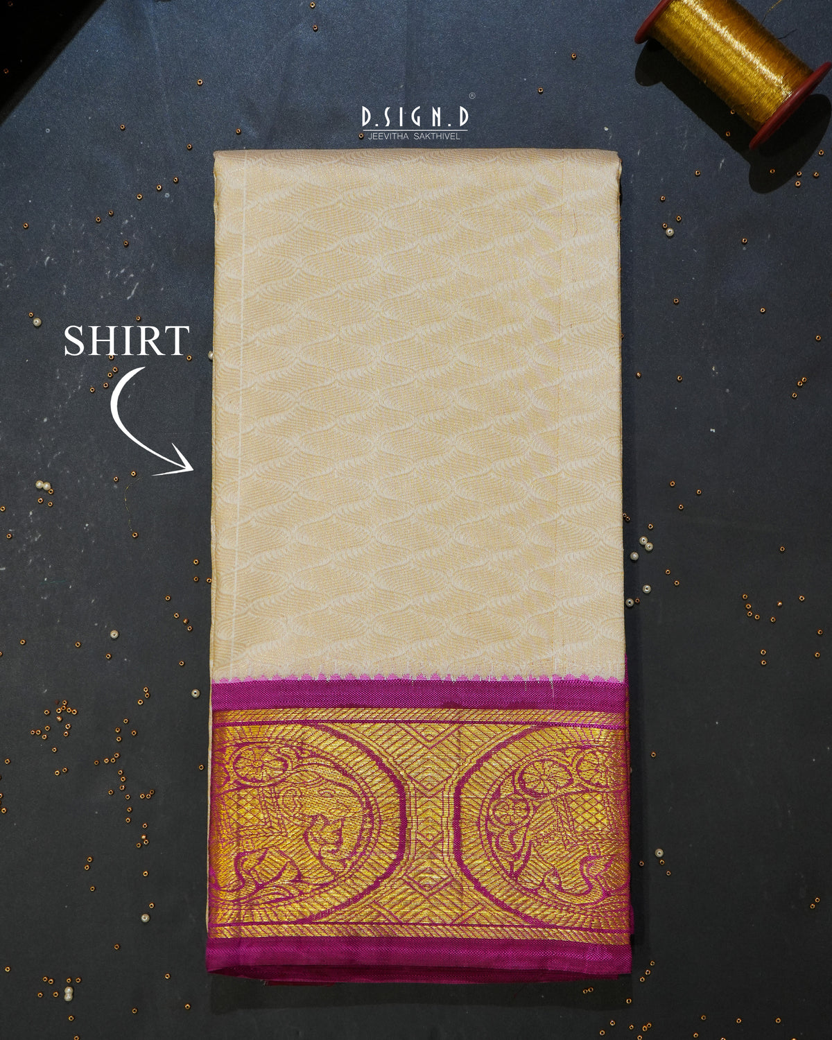 A folded cream-colored silk shirt material or fabric piece with a wide magenta and gold zari border, displayed on a dark textured surface with a spool of gold thread in the corner.