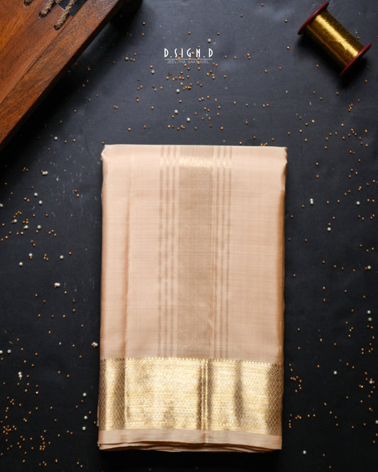 A folded beige silk dhoti with a wide metallic gold border, displayed on a dark textured background with decorative props.