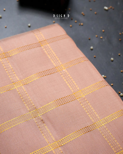 A close-up of a copper shade woven dhoti with a gold grid pattern, laid diagonally on a dark gray textured surface with scattered decorative beads.