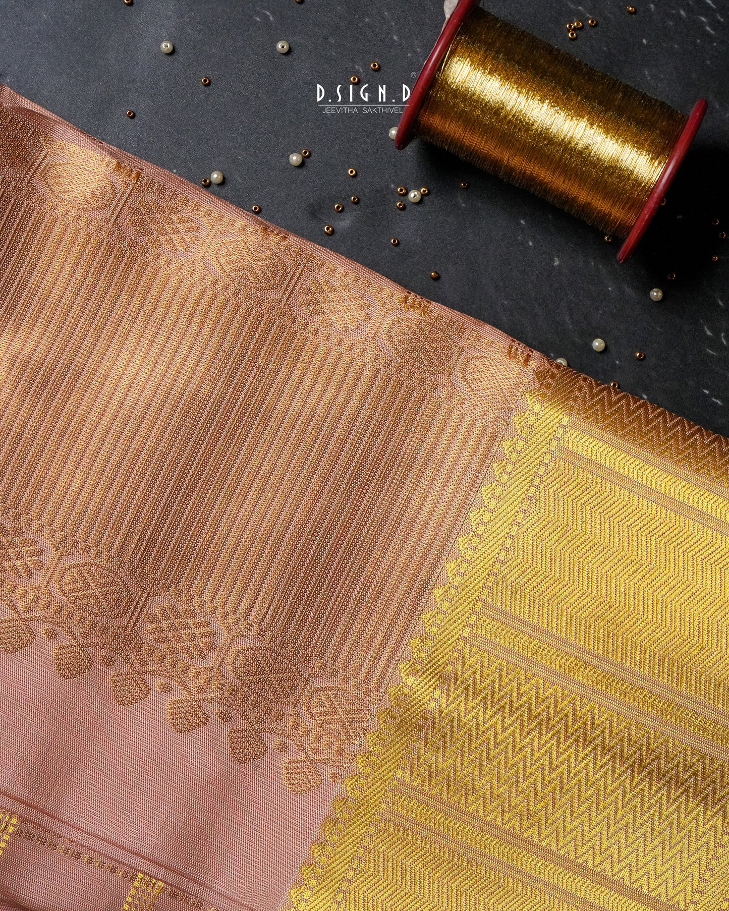 A close-up of copper and gold woven fabric with ornate metallic zari patterns, laid diagonally on a dark textured surface next to a spool of gold thread.