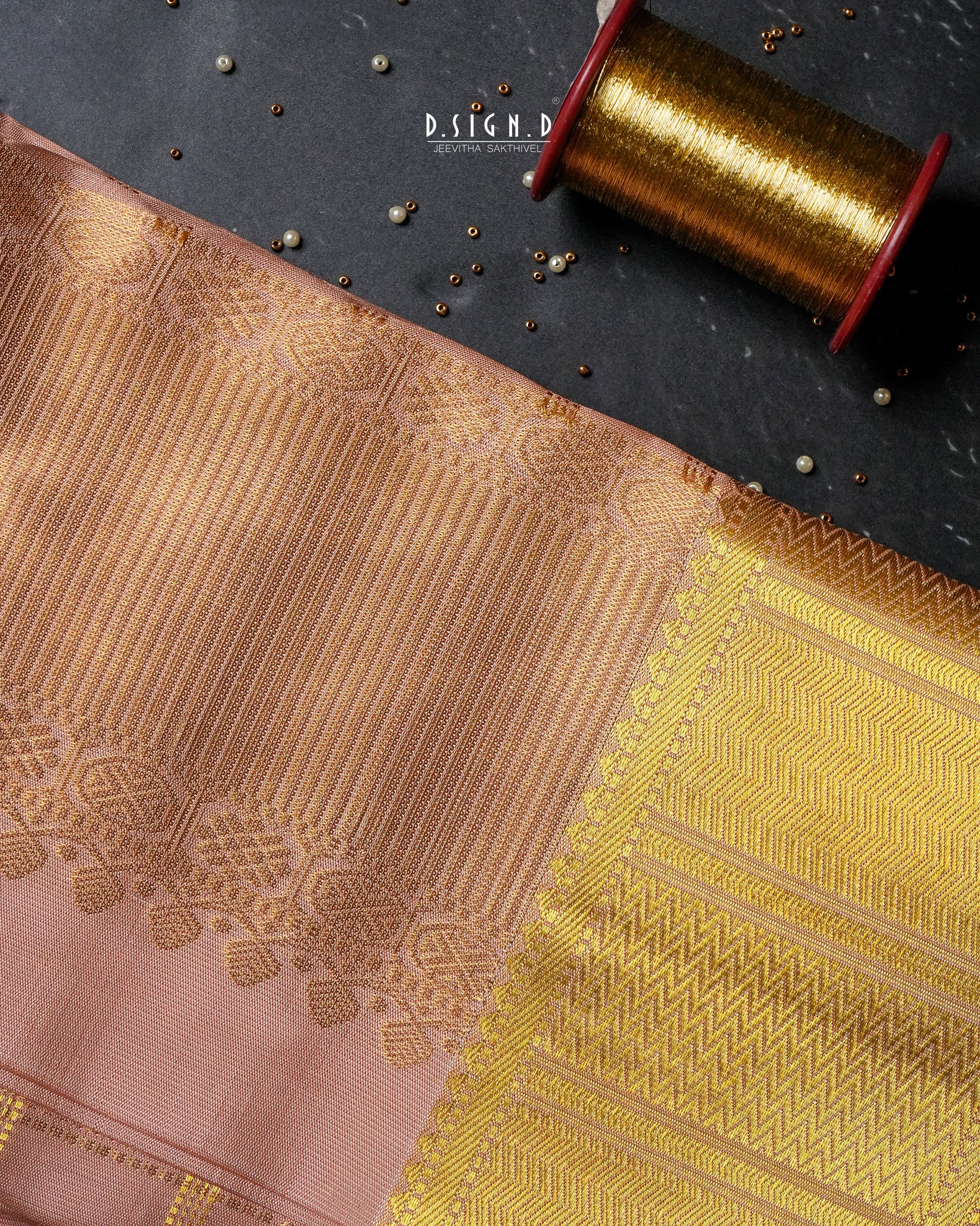 A close-up of copper and gold woven fabric with ornate metallic zari patterns, laid diagonally on a dark textured surface next to a spool of gold thread.