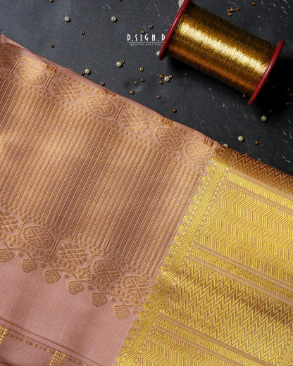 A close-up of copper and gold woven fabric with ornate metallic zari patterns, laid diagonally on a dark textured surface next to a spool of gold thread.