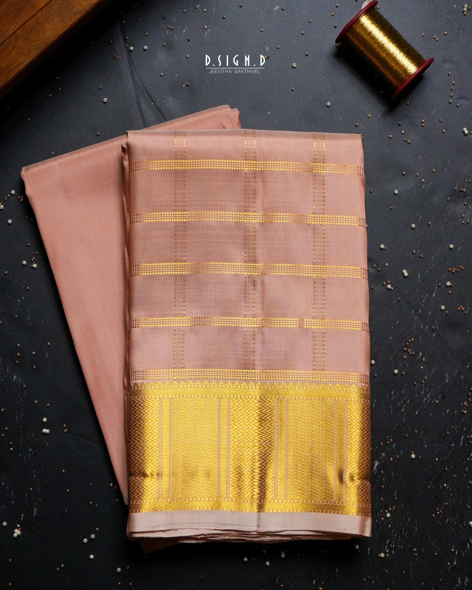 A folded copper dhothi set with a wide golden-yellow zari border, displayed on a dark textured background alongside a spool of gold thread.
