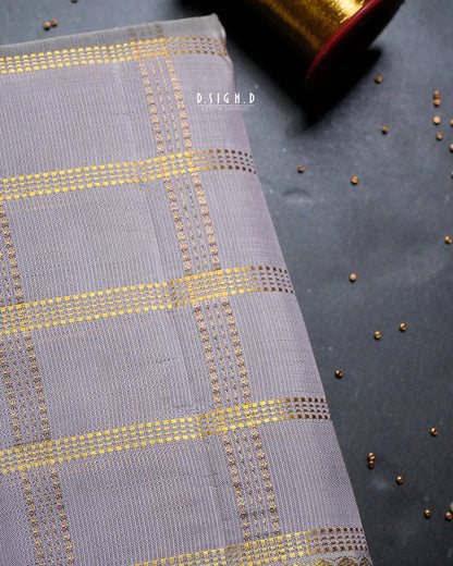 A close-up of a mauve dhoti with metallic gold woven checkered stripes, displayed on a dark textured surface with a spool of similar fabric in the background.