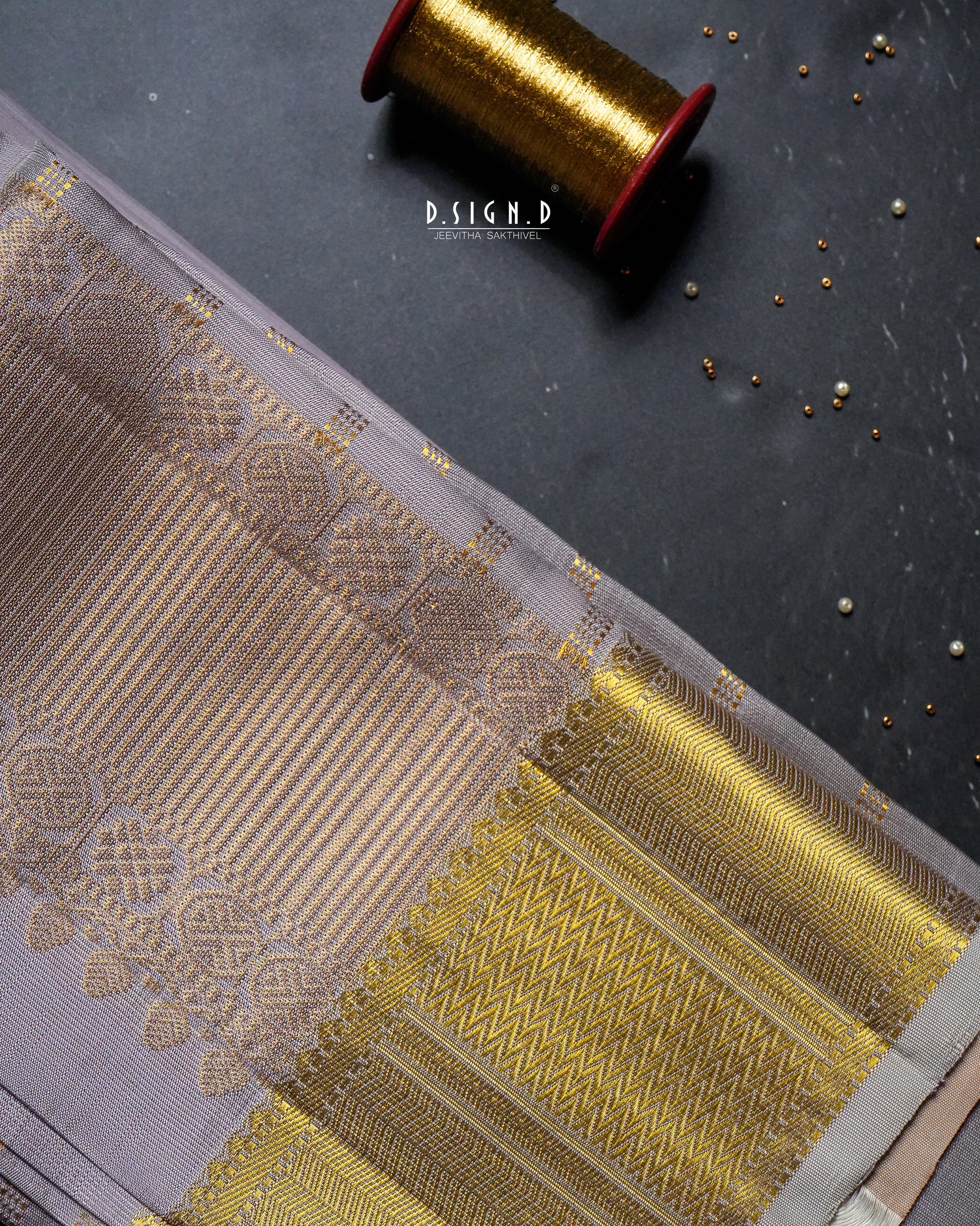 A close-up of a traditional Indian dhoti fabric with a mauve body and a wide gold zari border, laid diagonally on a dark textured surface with a spool of gold thread and small metallic embellishments.