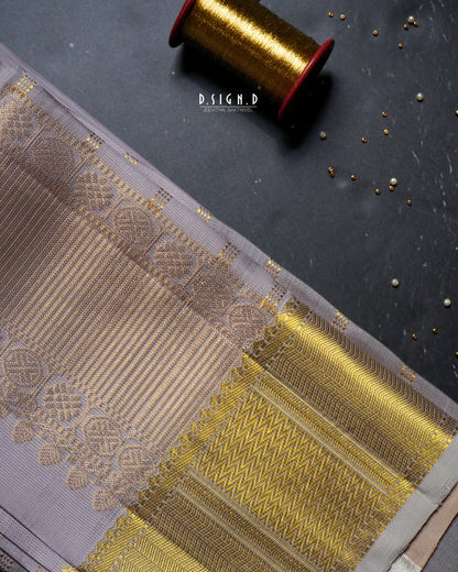 A close-up of a traditional Indian dhoti fabric with a mauve body and a wide gold zari border, laid diagonally on a dark textured surface with a spool of gold thread and small metallic embellishments.