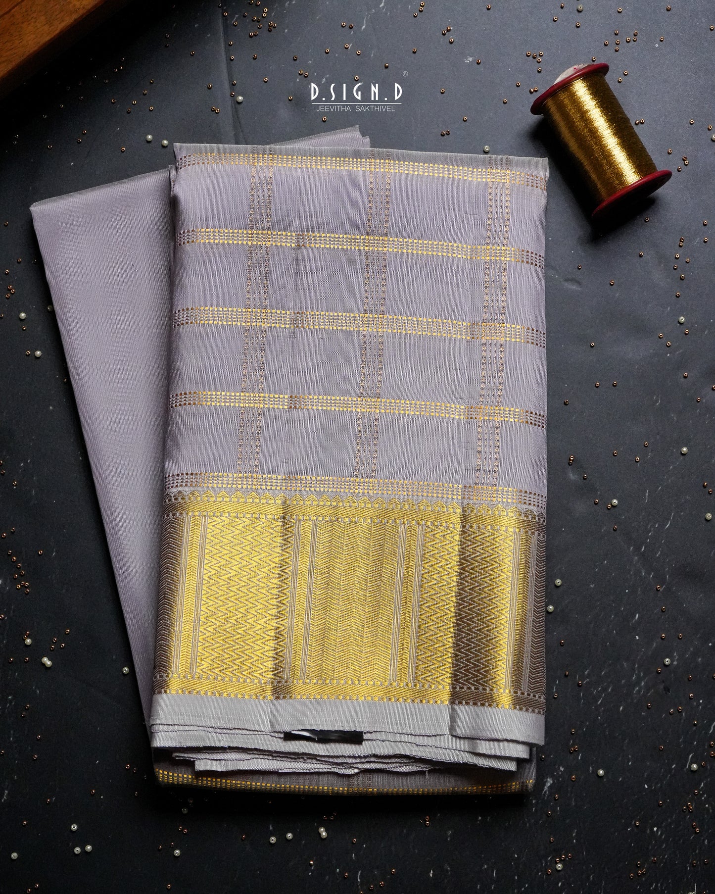 A folded mauve shade dhoti with gold woven stripes and a wide gold border & a plain shirt material , displayed on a dark textured surface next to a spool of gold thread. 