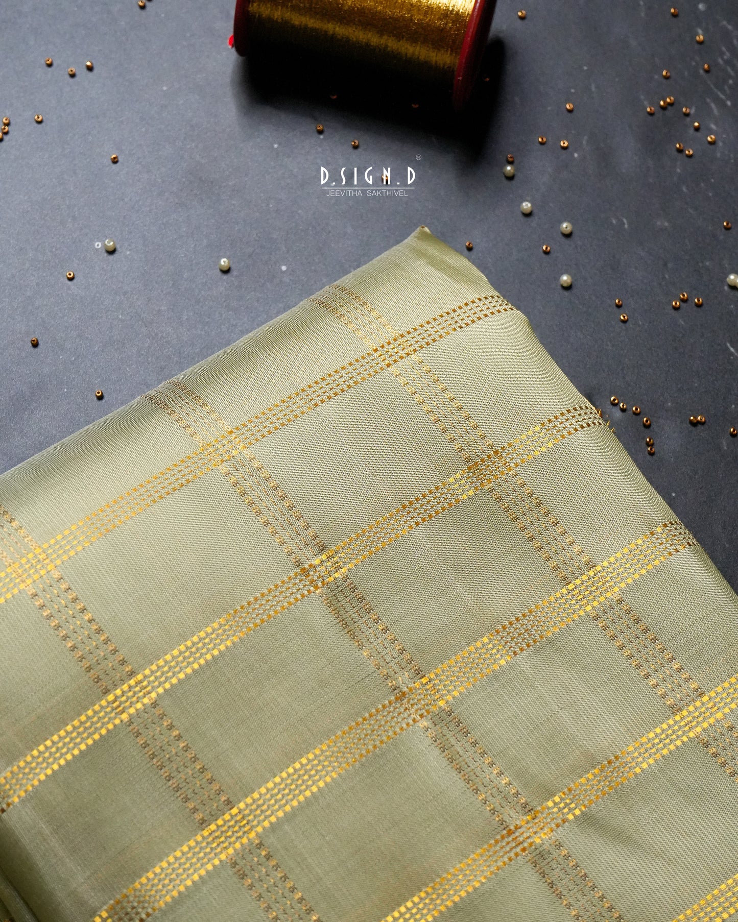 A folded piece of light pista green fabric with a gold woven checkered pattern, displayed on a dark gray textured surface with scattered metallic beads and a spool of gold thread in the background.