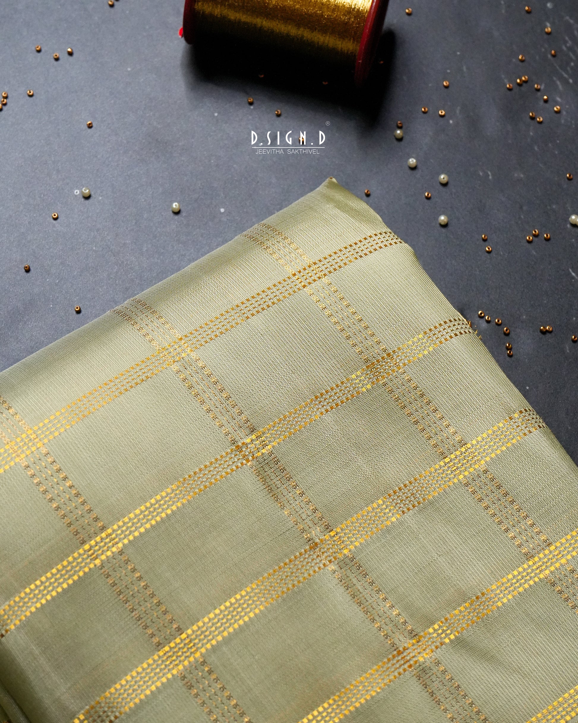 A folded piece of light pista green fabric with a gold woven checkered pattern, displayed on a dark gray textured surface with scattered metallic beads and a spool of gold thread in the background.