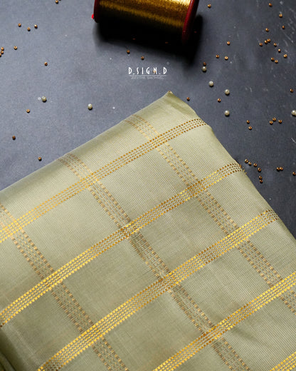 A folded piece of light pista green fabric with a gold woven checkered pattern, displayed on a dark gray textured surface with scattered metallic beads and a spool of gold thread in the background.