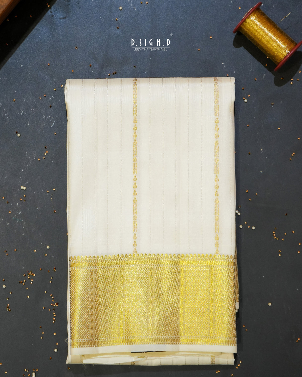 A folded white colored silk dhoti with a wide yellow and gold zari border, displayed on a dark textured surface with a spool of gold thread in the top right corner.