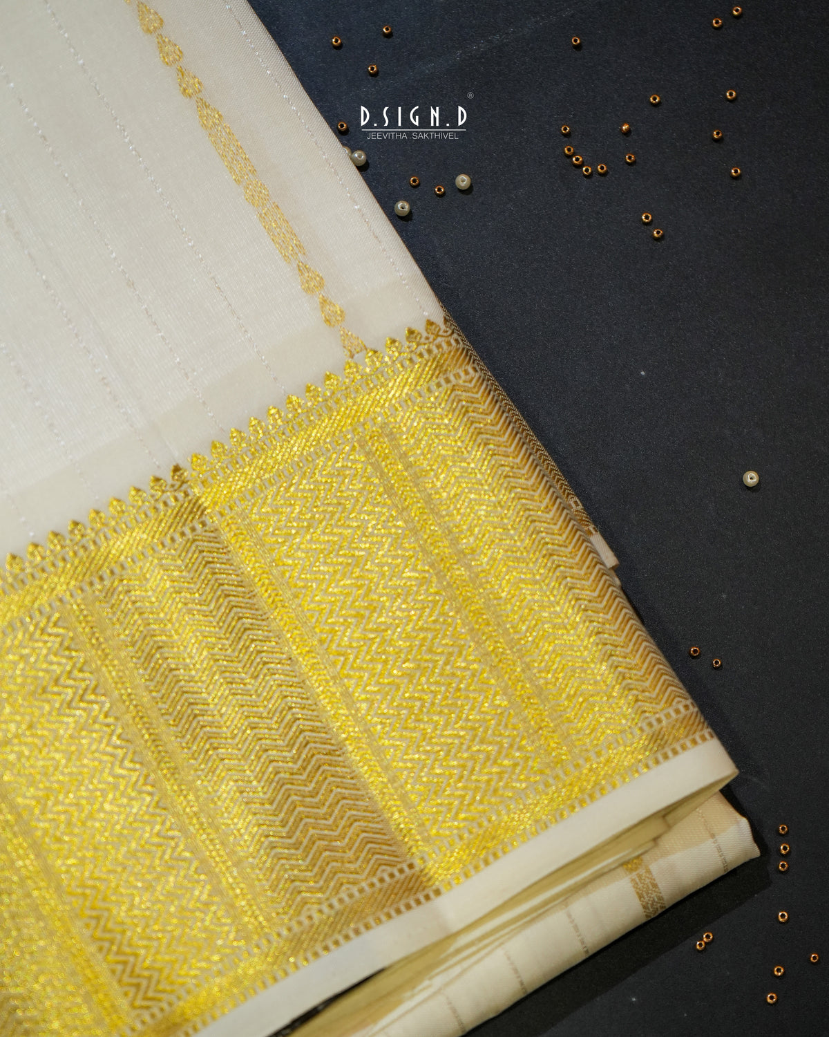 A folded white fabric, likely silk or satin, featuring an ornate golden-yellow & silver zari brocade border with a chevron and geometric pattern, displayed on a dark textured background with scattered metallic beads.
