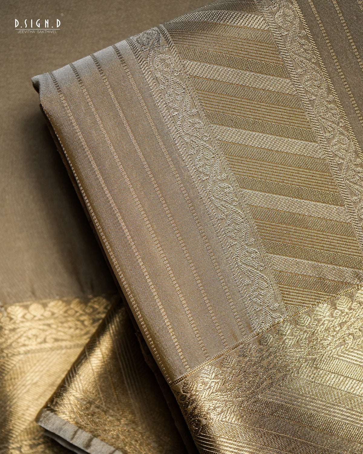 A close-up of a folded woven fabric, likely a textile, showing metallic gold and beige patterns with a subtle sheen. The fabric features vertical stripes, woven floral borders, and geometric designs.