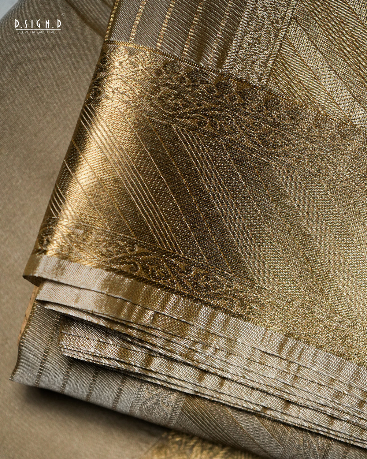 A close-up of folded brocade fabric in warm beige and metallic gold tones, showing woven patterns and sheen. The fabric fills the frame.