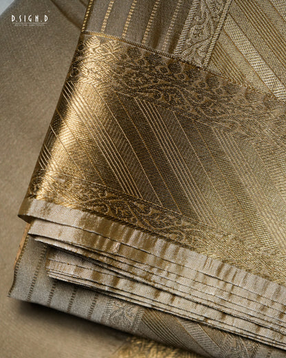 A close-up of folded brocade fabric in warm beige and metallic gold tones, showing woven patterns and sheen. The fabric fills the frame.