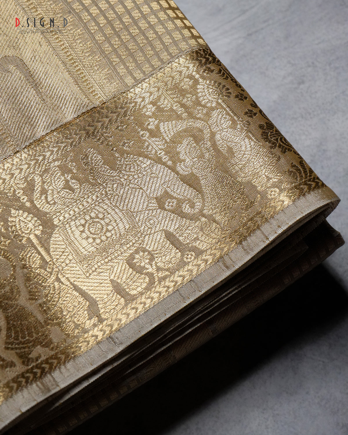 A folded gold and beige woven fabric, likely a silk dhoti or textile, featuring an intricate elephant and floral motif in the border, displayed on a textured gray surface.