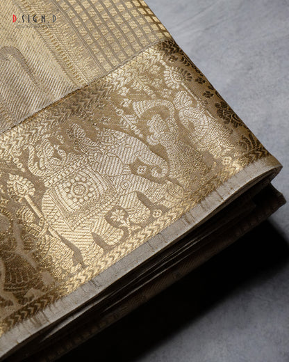 A folded gold and beige woven fabric, likely a silk dhoti or textile, featuring an intricate elephant and floral motif in the border, displayed on a textured gray surface.