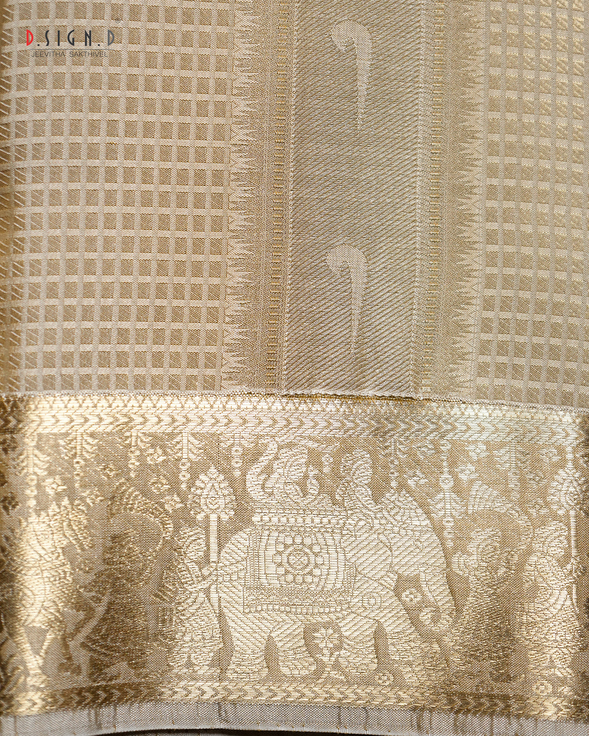 A close-up of a beige and gold woven fabric, likely a dhoti body or decorative textile, featuring a detailed gold zari brocade border with traditional motifs and a grid pattern.