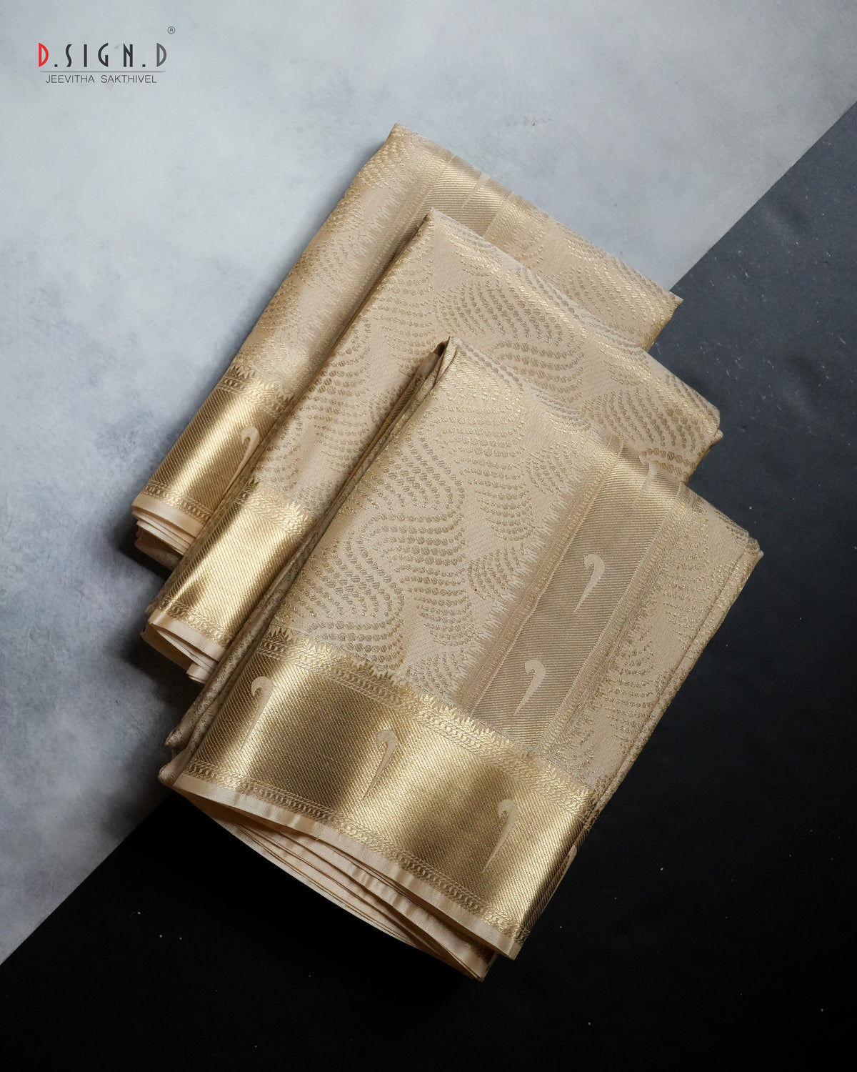 A folded gold silk dhoti set with a metallic gold border and subtle woven patterns, displayed on a split gray and black background. 