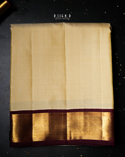 A folded cream-colored dhoti with a wide metallic gold border and a deep maroon pallu, displayed flat on a dark textured background with gold glitter.