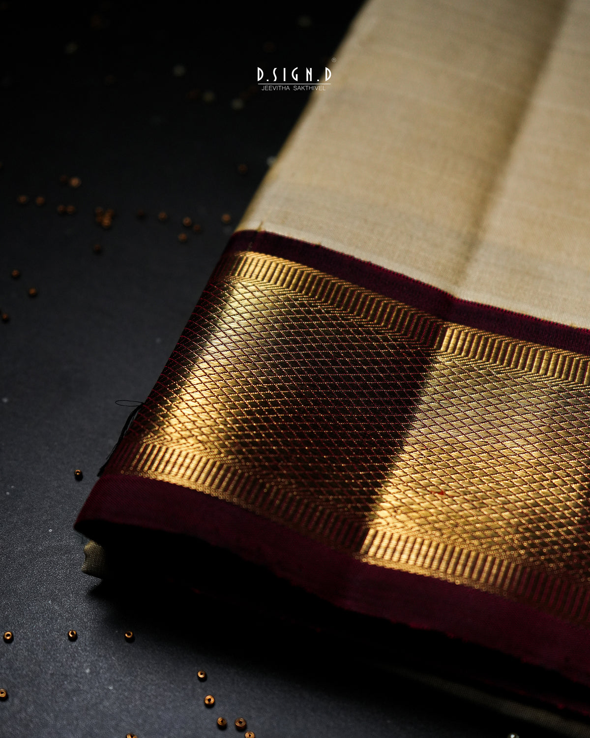 A close-up of a traditional Indian dhoti fabric with a beige body, a wide metallic gold zari border, and a deep maroon pallu, displayed on a dark textured surface.