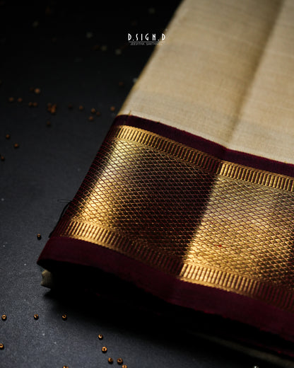 A close-up of a traditional Indian dhoti fabric with a beige body, a wide metallic gold zari border, and a deep maroon pallu, displayed on a dark textured surface.