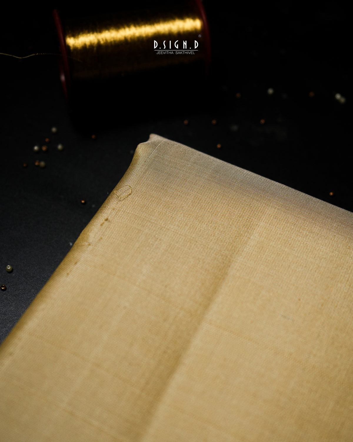 A close-up of a folded beige fabric with a visible woven texture, set against a dark background with a blurred gold spool in the upper corner. 