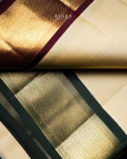 A close-up of a folded silk dhoti or fabric, showing a cream-colored body with a wide gold zari border and a dark green and maroon pallu.