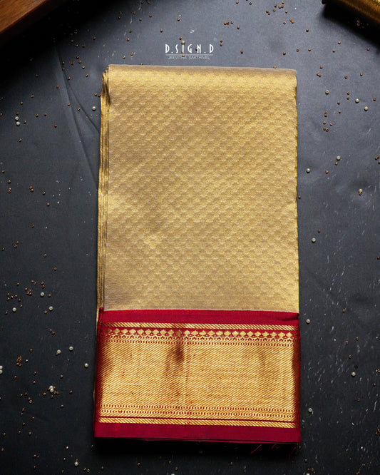 A folded golden tissue pure silk shirt material with a red border and intricate gold zari work , displayed flat on a dark textured surface .