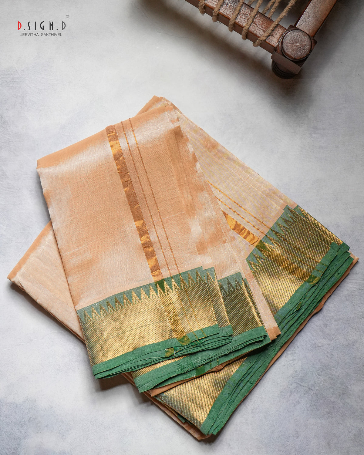 A folded peach-colored silk dhoti & vastram with a wide gold zari border and a green pallu, displayed on a textured gray surface with a wooden object partially visible in the top right corner.