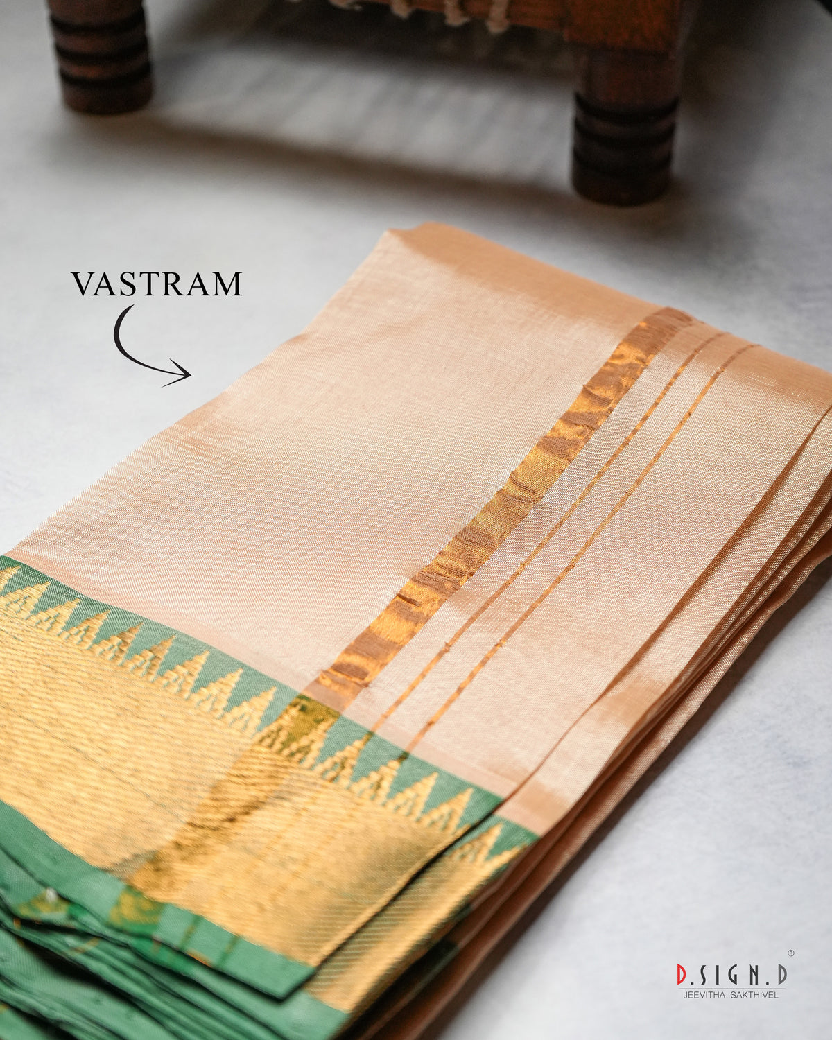 A folded beige silk vastram with a green and gold zari border. The fabric is displayed on a light gray surface with a wooden leg visible in the background.