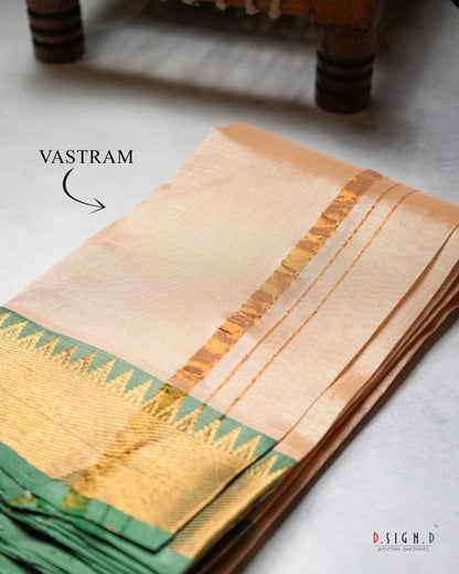 A folded beige silk vastram with a green and gold zari border. The fabric is displayed on a light gray surface with a wooden leg visible in the background.