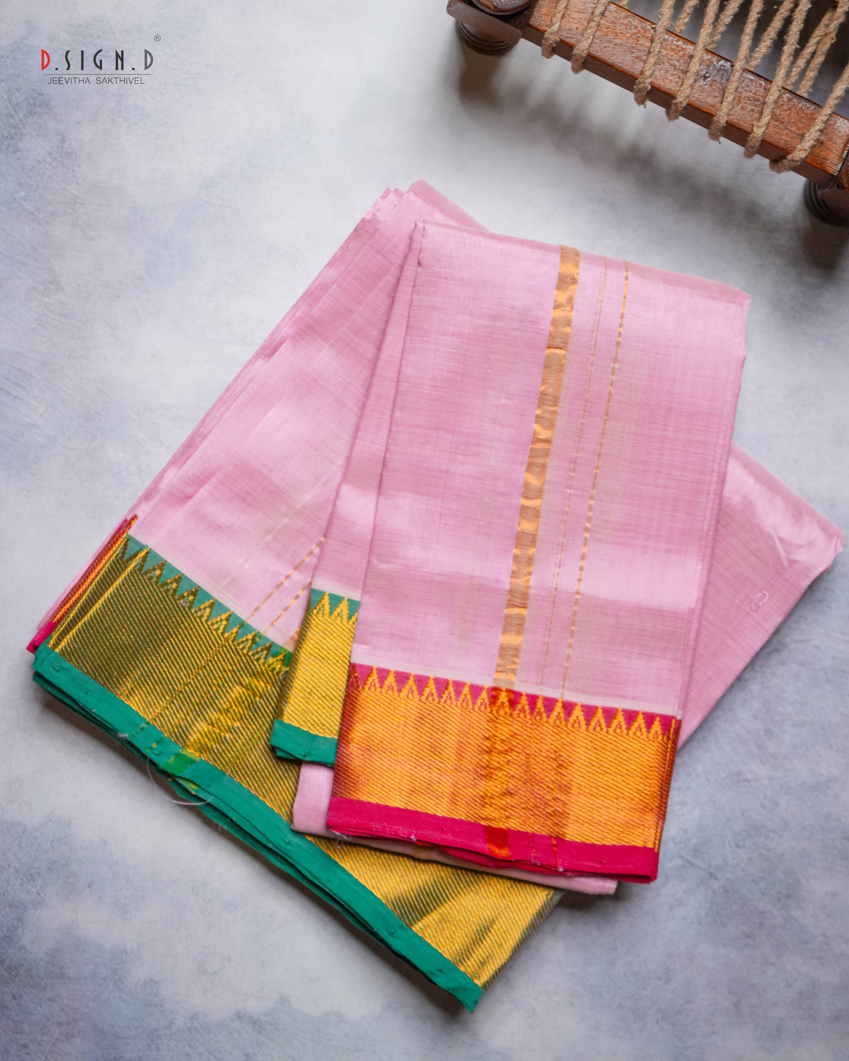 A folded pink silk Dhoti with a wide gold zari border and a contrasting green and pink pallu, displayed on a textured gray surface.