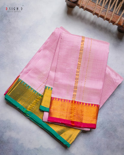 A folded pink silk Dhoti with a wide gold zari border and a contrasting green and pink pallu, displayed on a textured gray surface.