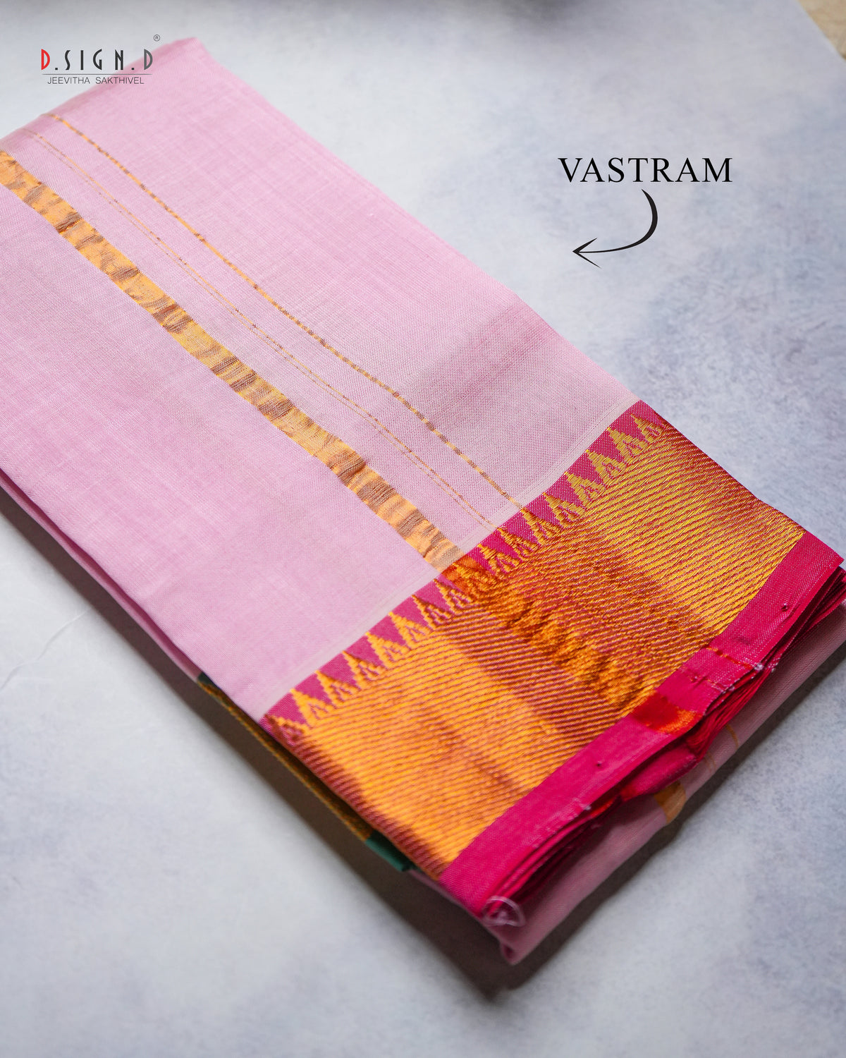 A folded pink silk vastram with a wide golden-yellow and magenta border, displayed on a textured gray surface. 