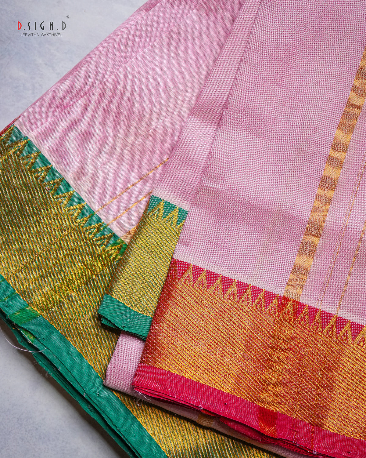 A folded pink silk dhoti & vastram with a wide golden-yellow zari border, featuring green and pink geometric accents, displayed on a light gray surface.