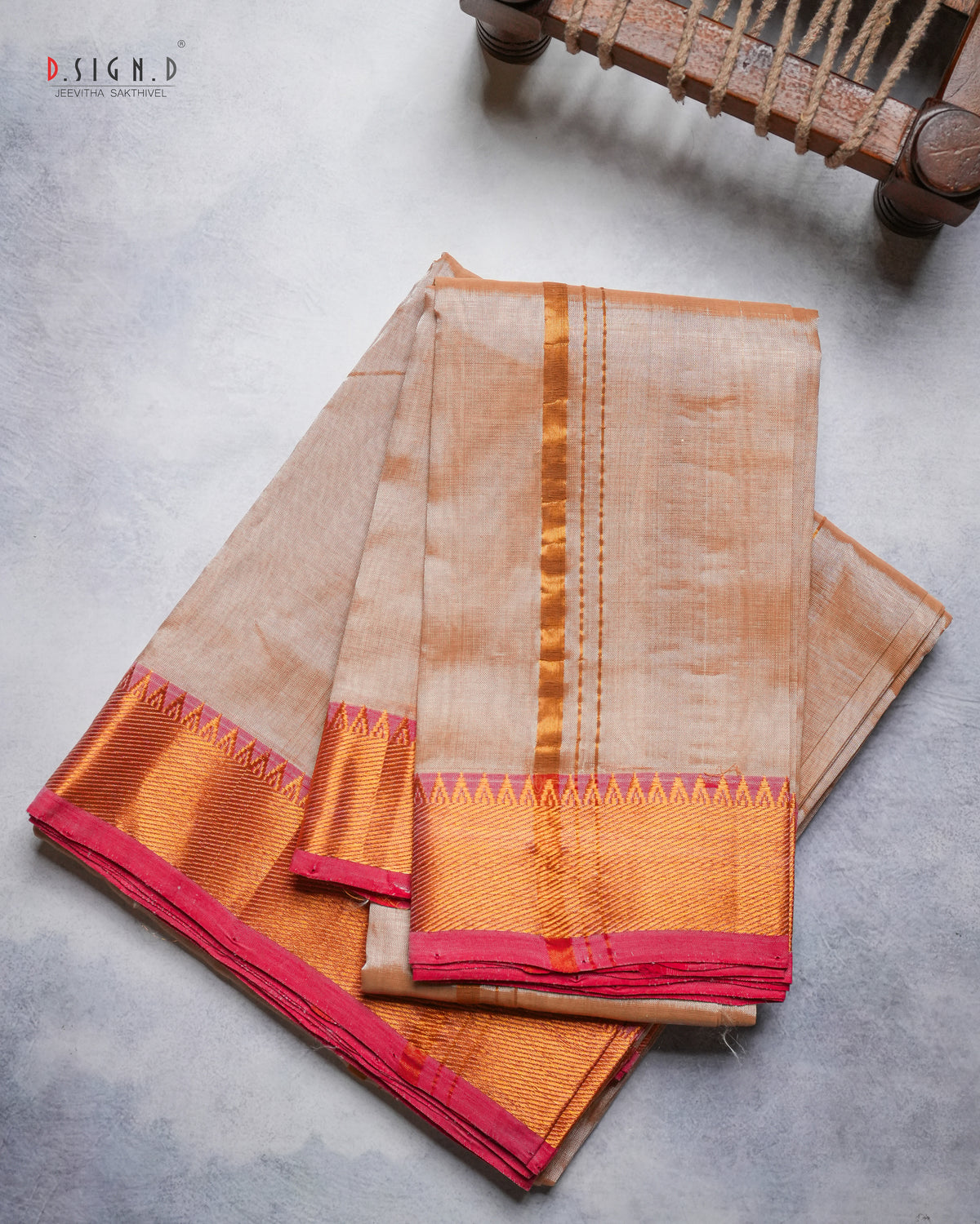 A folded copper silk dhoti & vastram with a vibrant red border, displayed on a textured gray surface with a wooden chair leg visible in the top right corner.