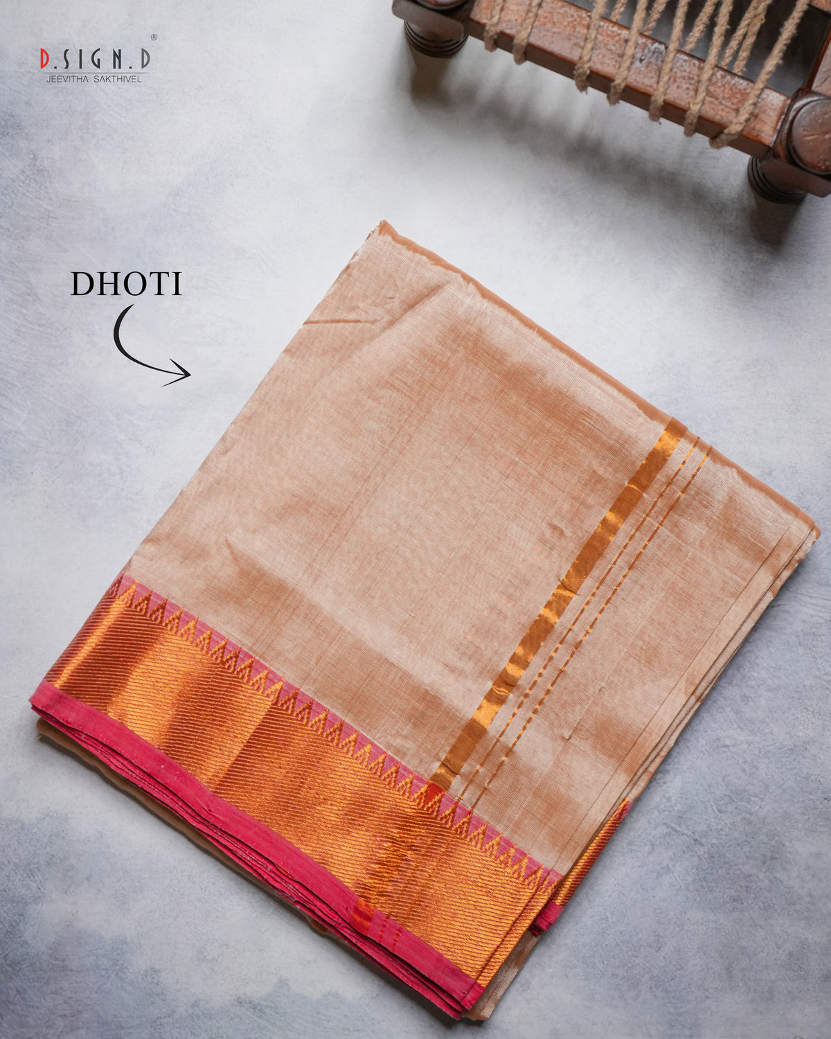 A folded Dhoti from Design D, displayed on a textured gray surface. The fabric is a warm copper color with a vibrant red border, and a gold zari stripe.