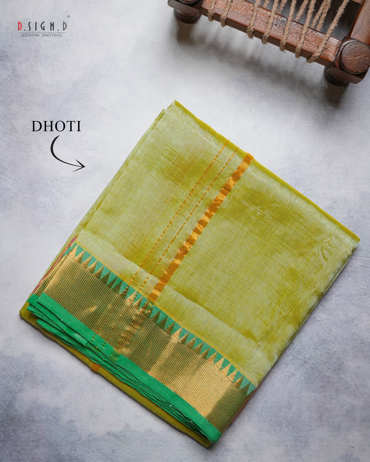 A folded yellow-green Dhoti with a gold and green border, displayed on a textured gray surface with a wooden loom visible in the top right corner.