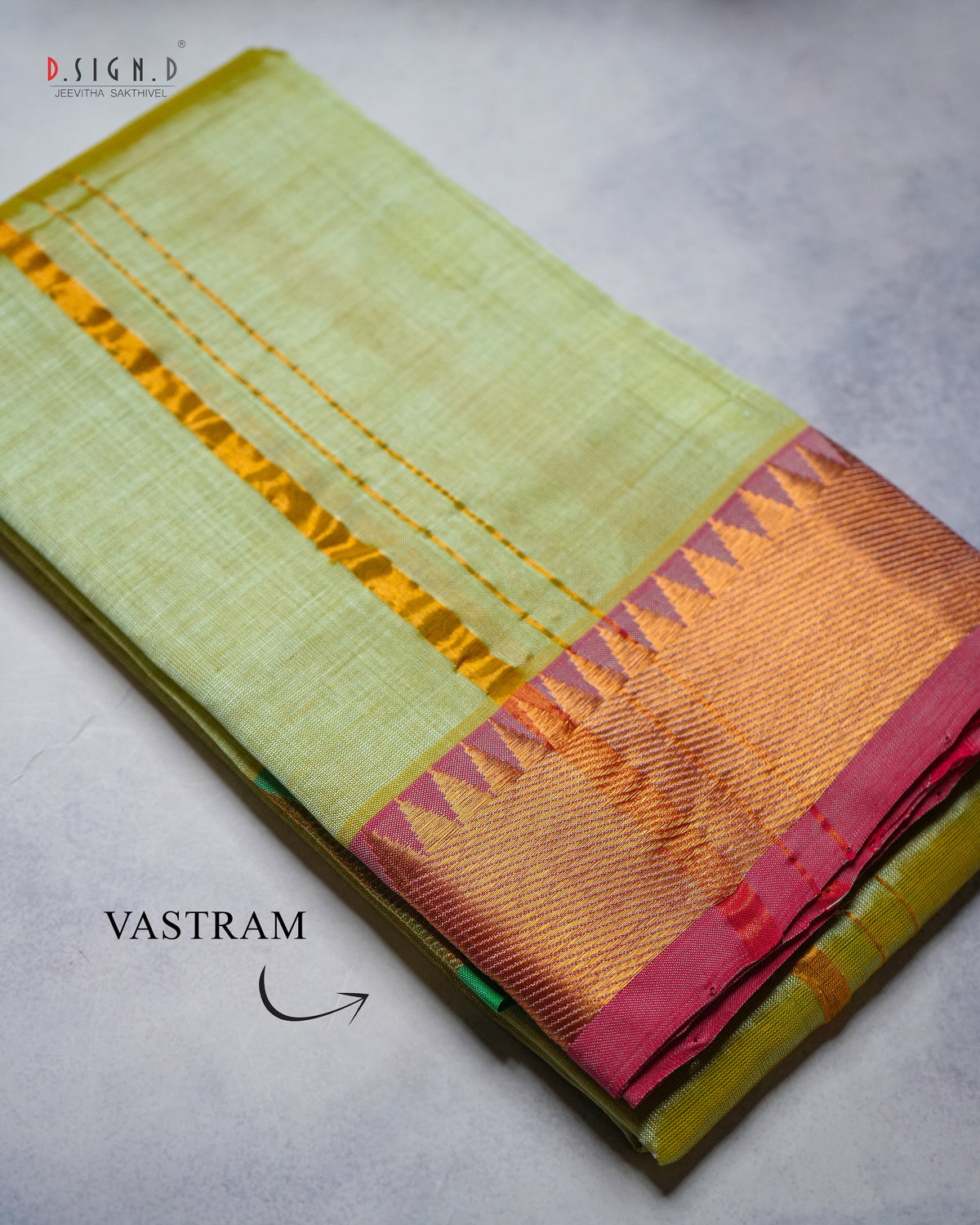 A folded light green silk vastram with a wide golden-yellow Vastram border and a magenta-pink and gold zari accent, displayed on a textured gray surface.