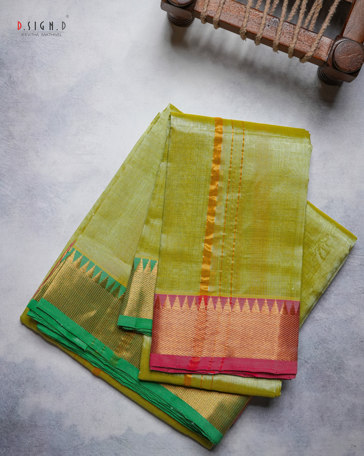 A folded olive green silk dhoti & vastram with a gold zari border and a pink and green pallu, displayed on a textured gray surface with a wooden chair leg visible in the top right corner.