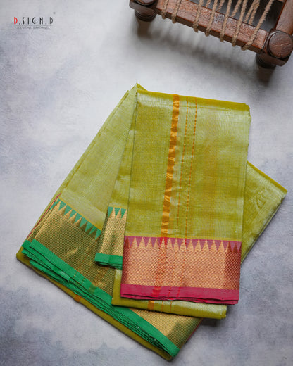 A folded olive green silk dhoti & vastram with a gold zari border and a pink and green pallu, displayed on a textured gray surface with a wooden chair leg visible in the top right corner.