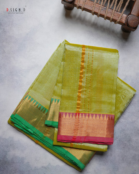 A folded olive green silk dhoti & vastram with a gold zari border and a pink and green pallu, displayed on a textured gray surface with a wooden chair leg visible in the top right corner.