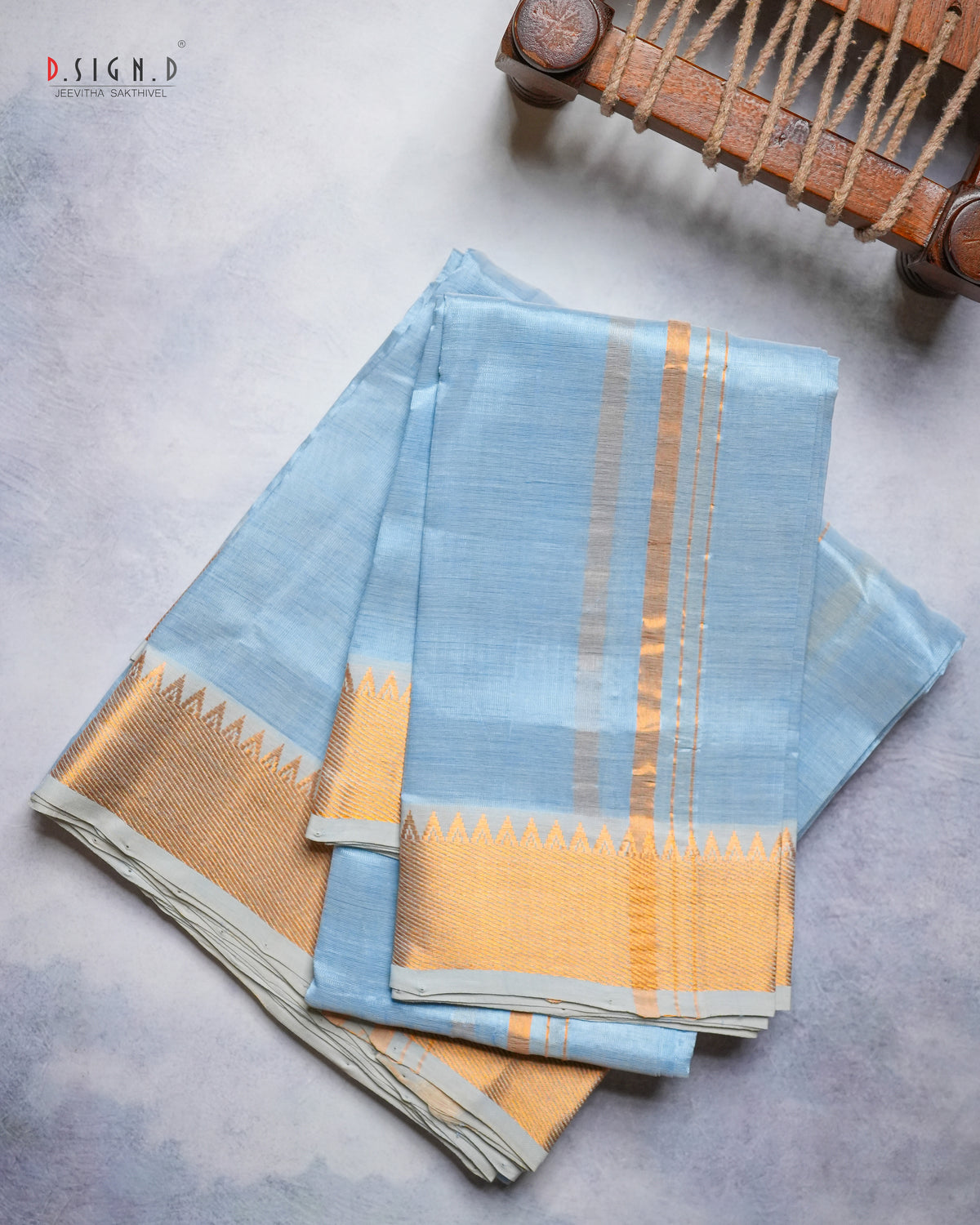 A folded light blue silk dhoti & vastram with a wide gold zari border and vertical gold stripes, displayed on a textured gray surface with a wooden loom visible in the top right corner.